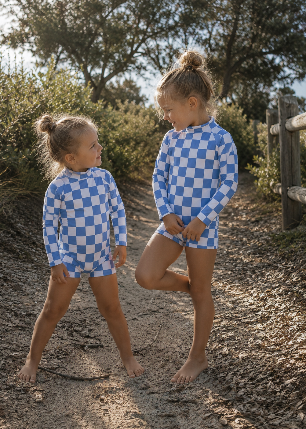 Two young girls standing barefoot on a dirt path outdoors, wearing matching blue and white checkered long-sleeve swim shirts and shorts.