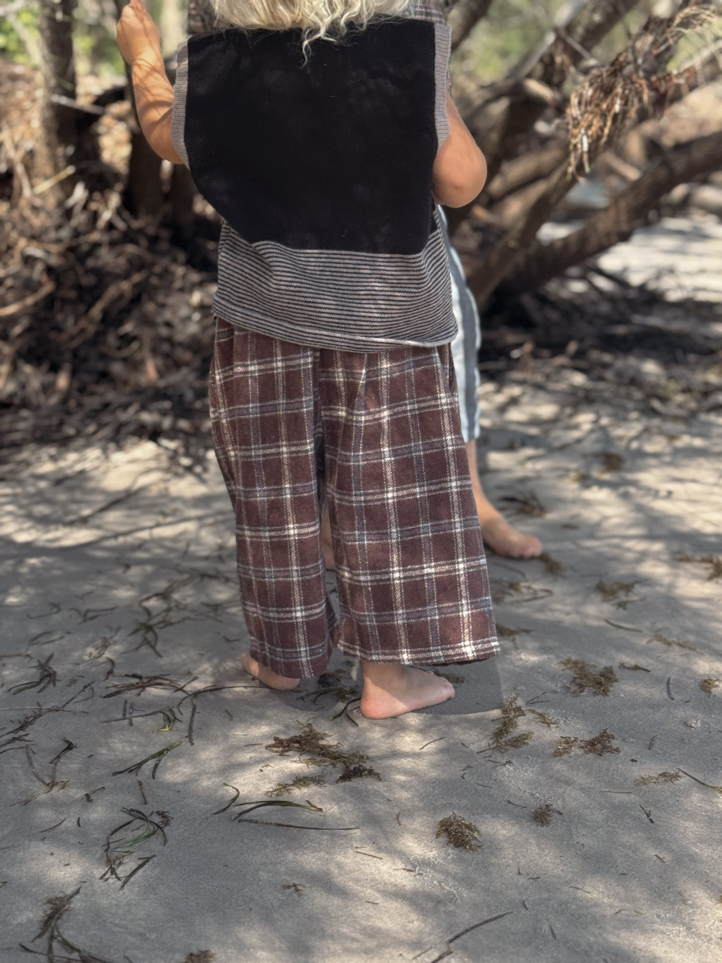Person wearing plaid pants and a black top standing on a sandy beach with seaweed.