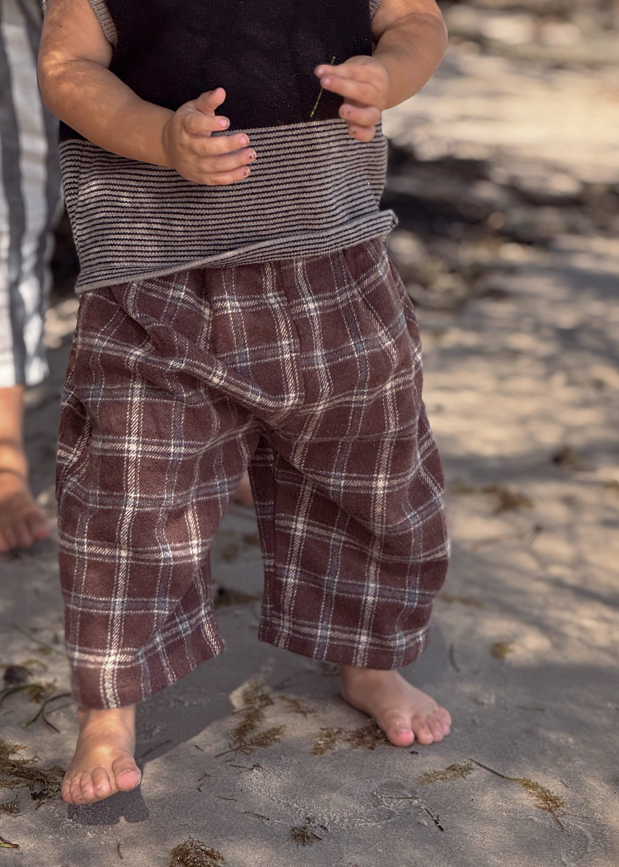 Child wearing a plaid outfit standing on a sandy beach with another person partially visible.