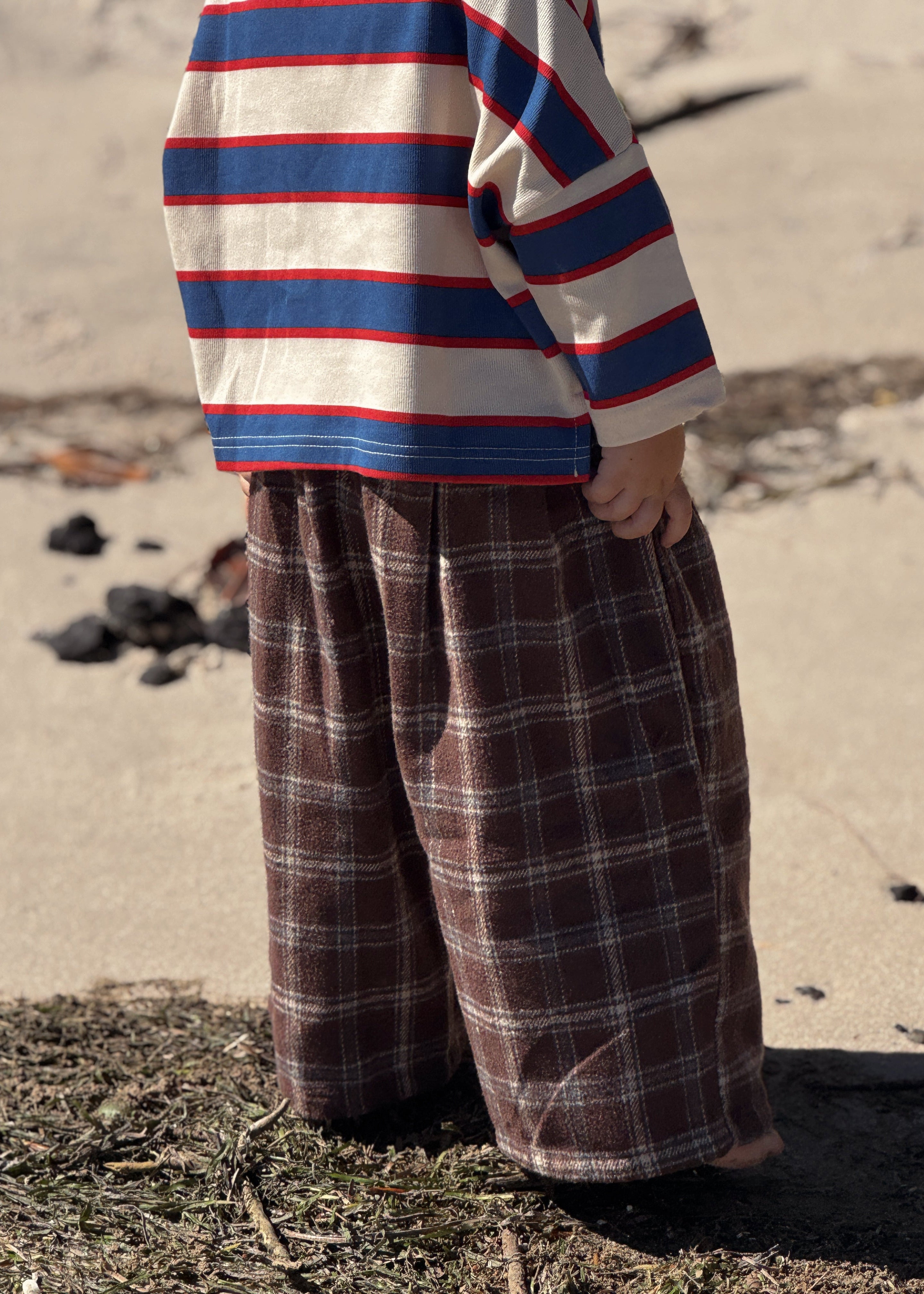 A child standing outdoors on sand and dark ground, wearing a cream long-sleeve shirt with blue and red horizontal stripes and wide brown plaid pants.