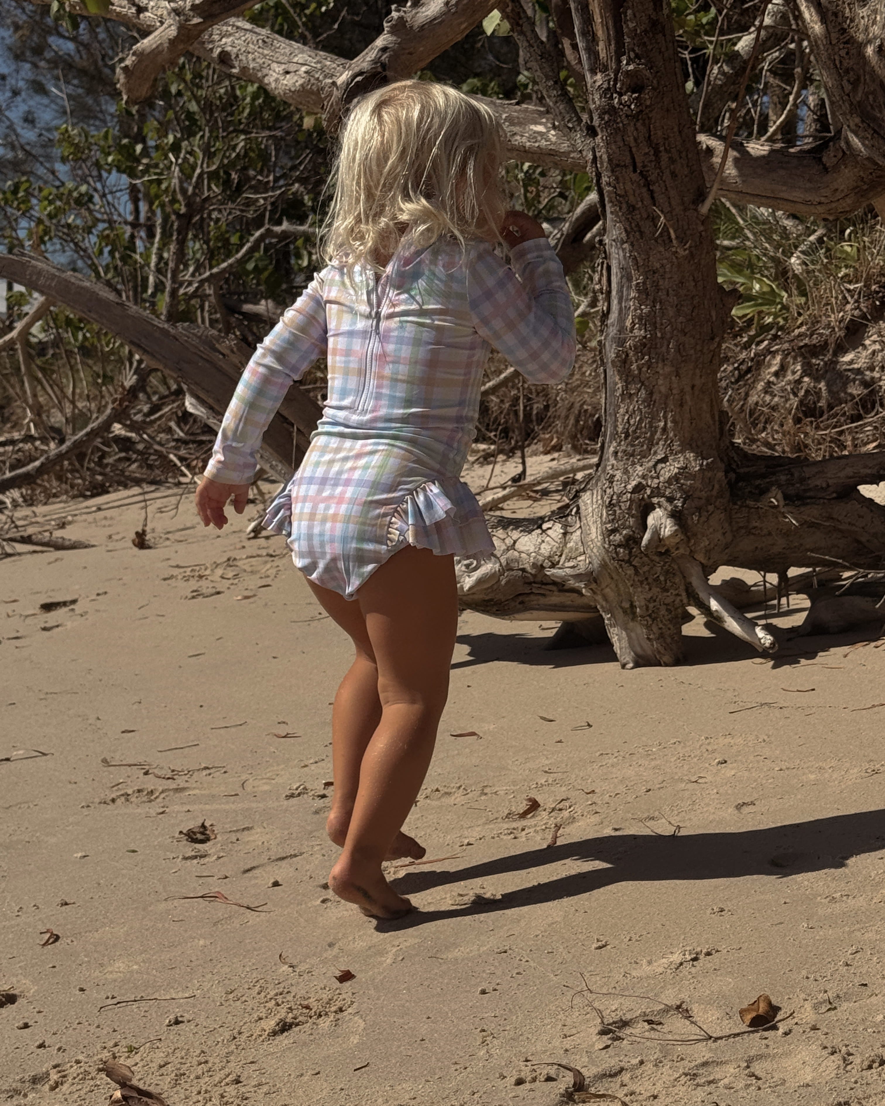 Child in a bright sun safe gingham swimsuit with snaps running on a sandy beach with trees in the background