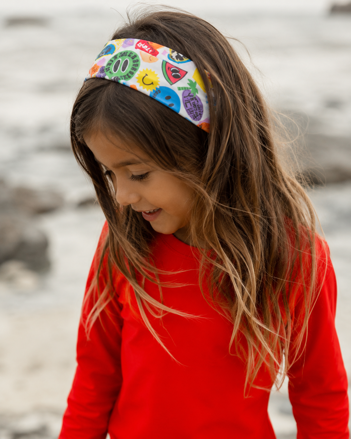 Child wearing a colorful headband with a pattern of various symbols, standing by a body of water.