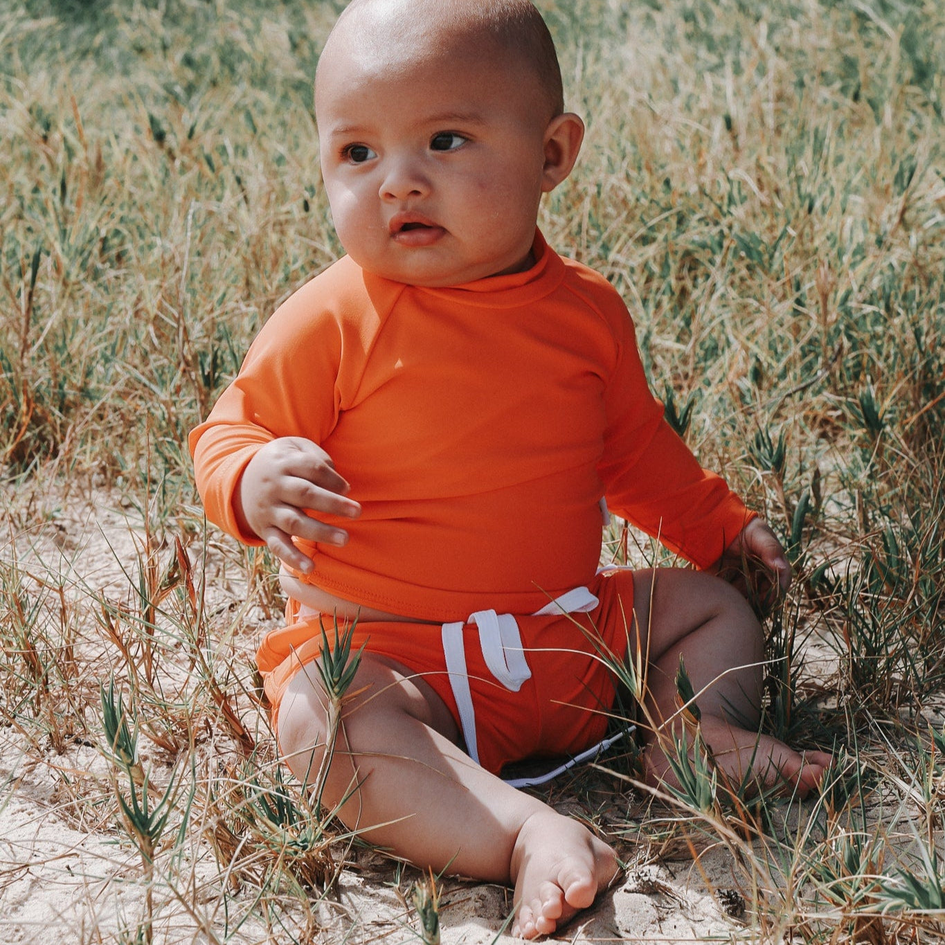 Baby in an bright orange swim shirt and swim short sitting in a sandy field