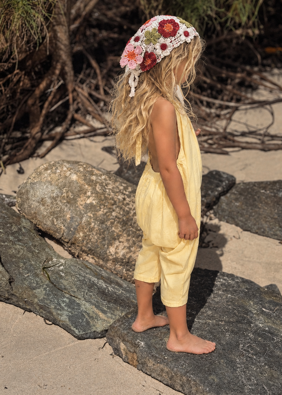A young child standing barefoot on large rocks on a sandy beach, wearing a pale yellow sleeveless romper and a white crochet sun hat with red and green floral embroidery.