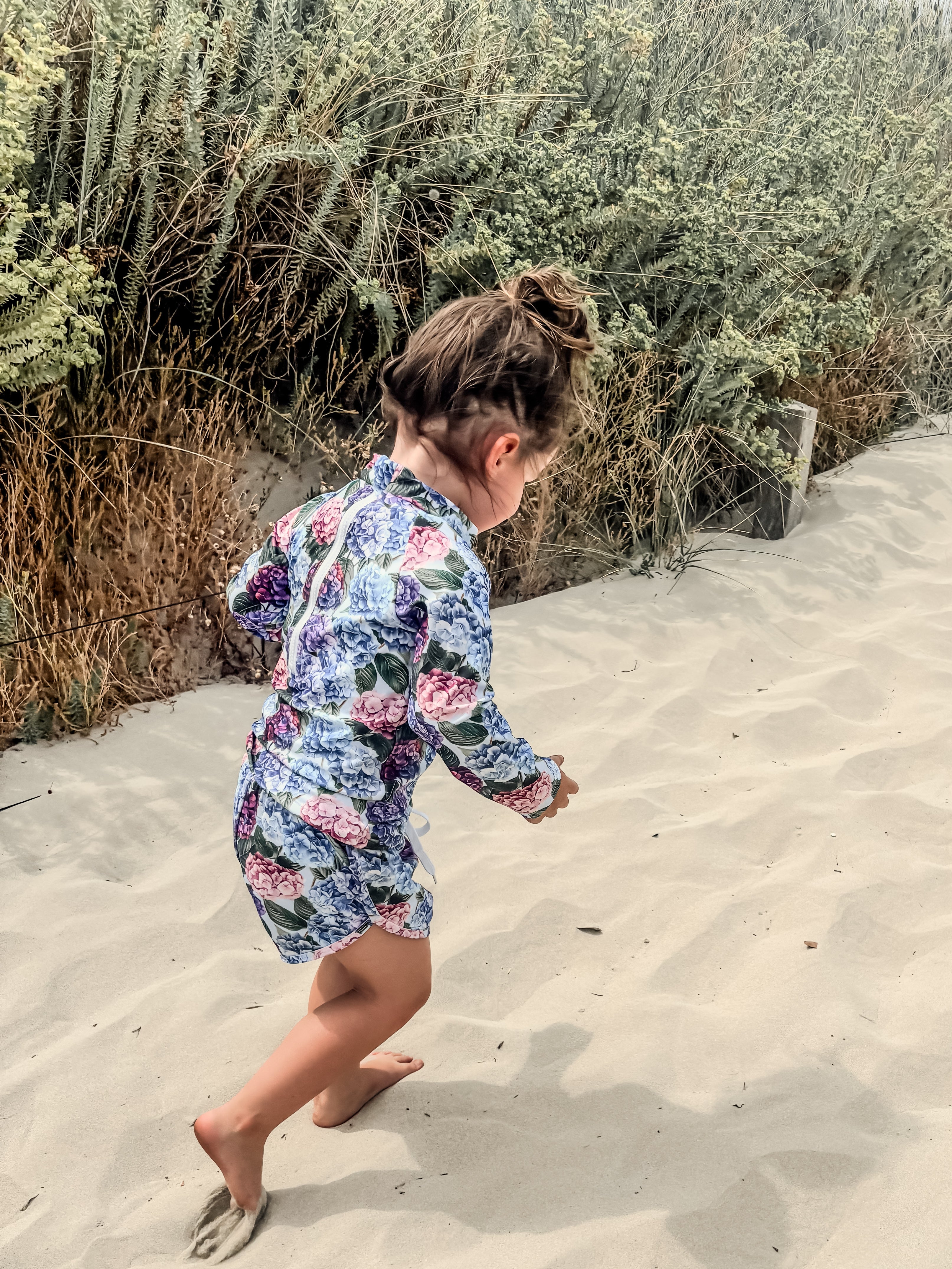toddler girl in a floral rash set walking on sand with plants in the background