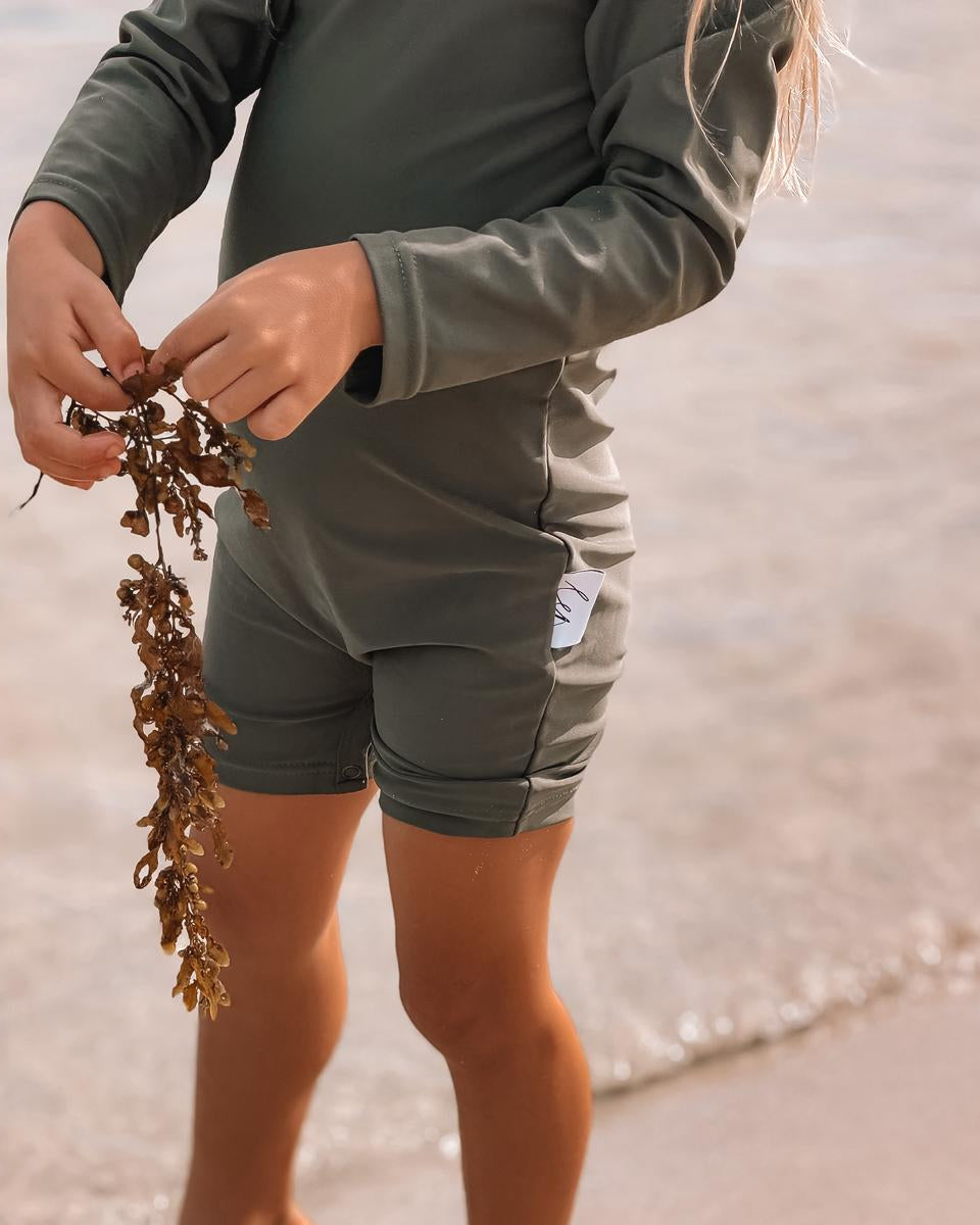 Person wearing a green outfit standing on a beach holding seaweed.