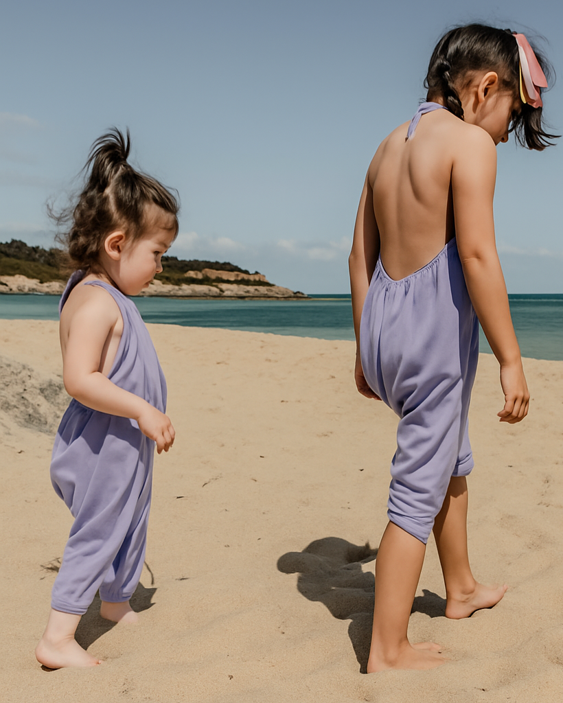 Two children in purple rompers standing on a sandy beach with clear blue sky.