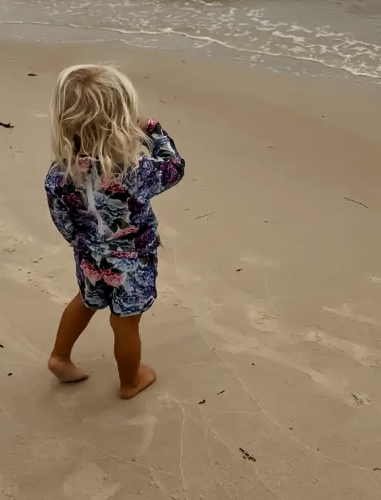 Child in a floral girls rash set  outfit standing on a sandy beach with waves in the background