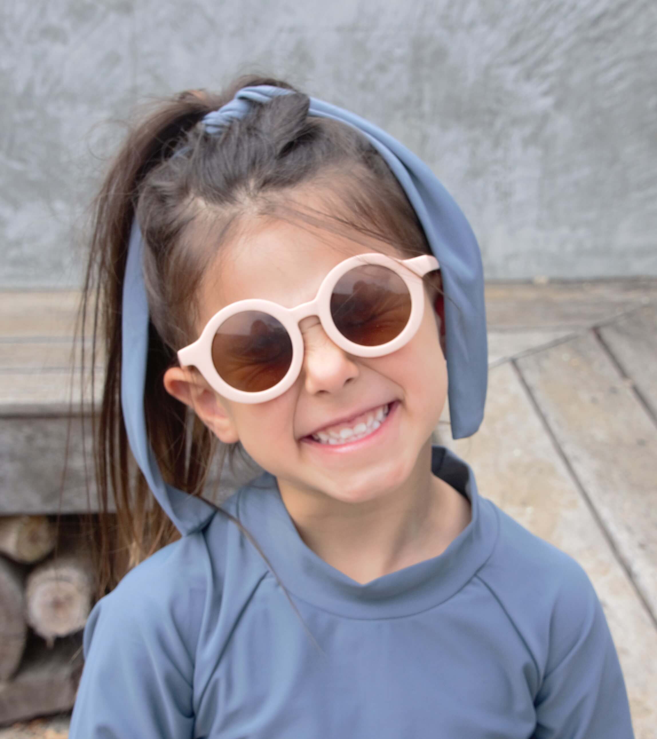 girl looking at the camera, wearing round pink sunglasses and blue rash top with matching headband, smiling