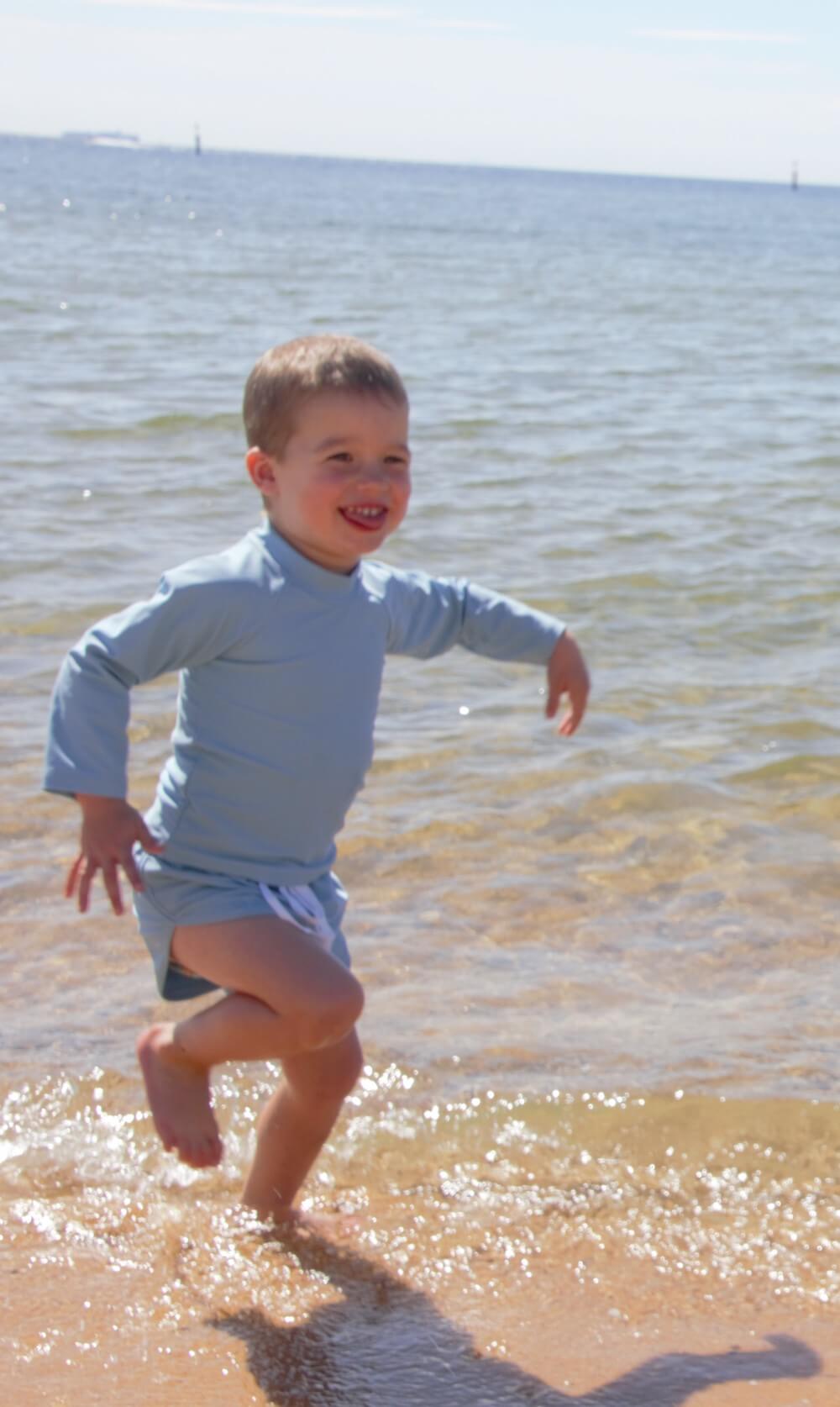 a boy running in the water on the beach smiling, wearing a blue rash set