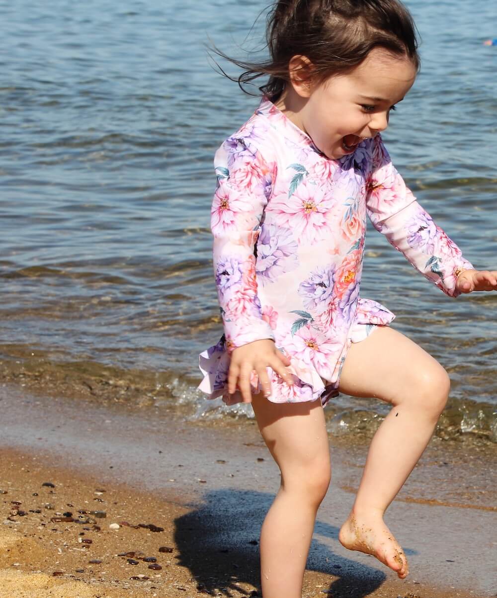 a girl playing and smiling on the beach near the water wearing long sleeve floral swimsuit with playful frills