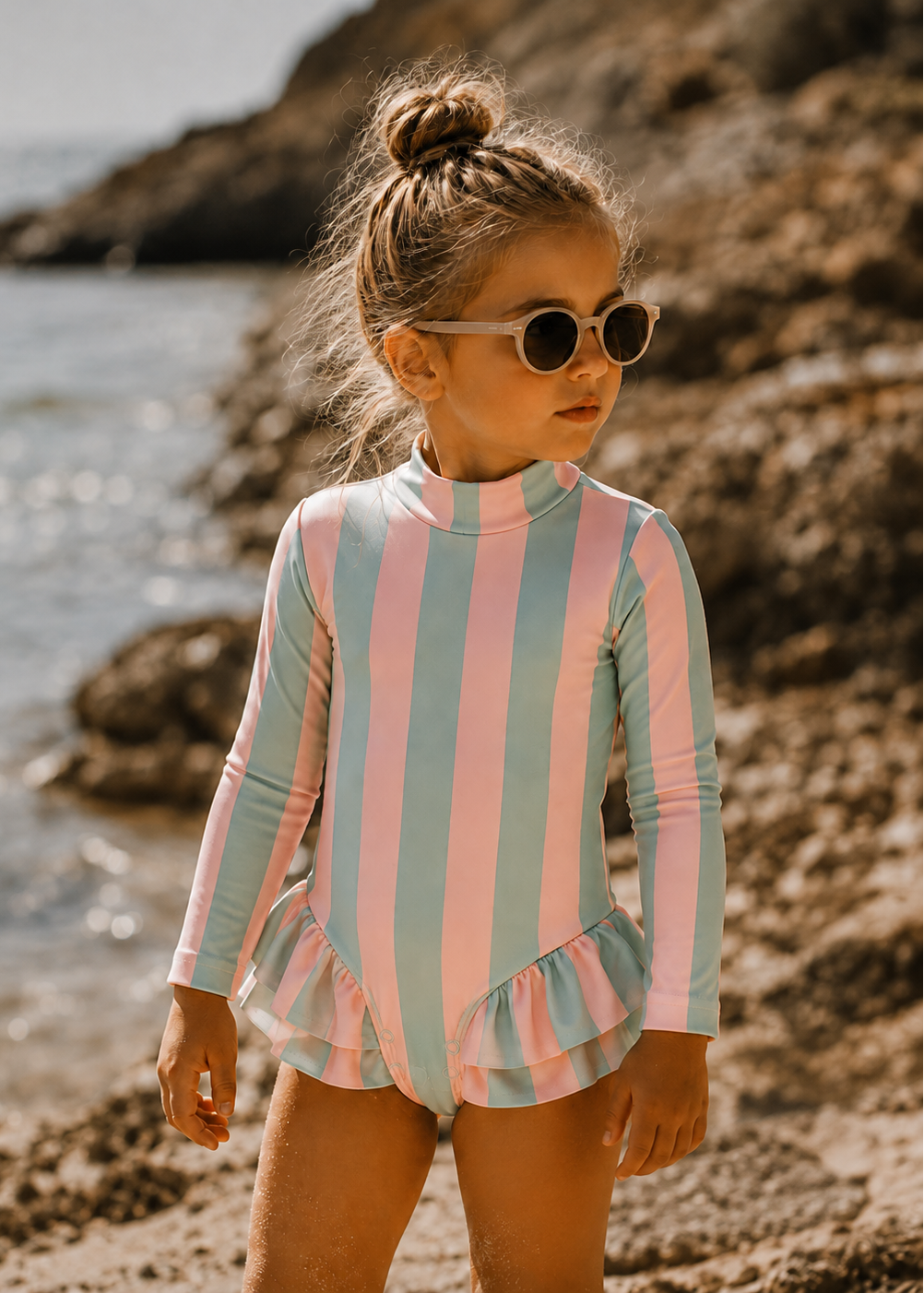 A young girl with her hair in a bun and wearing round sunglasses stands on a rocky beach, wearing a long-sleeve swim shirt with vertical pink and light blue stripes and a ruffled hem.
