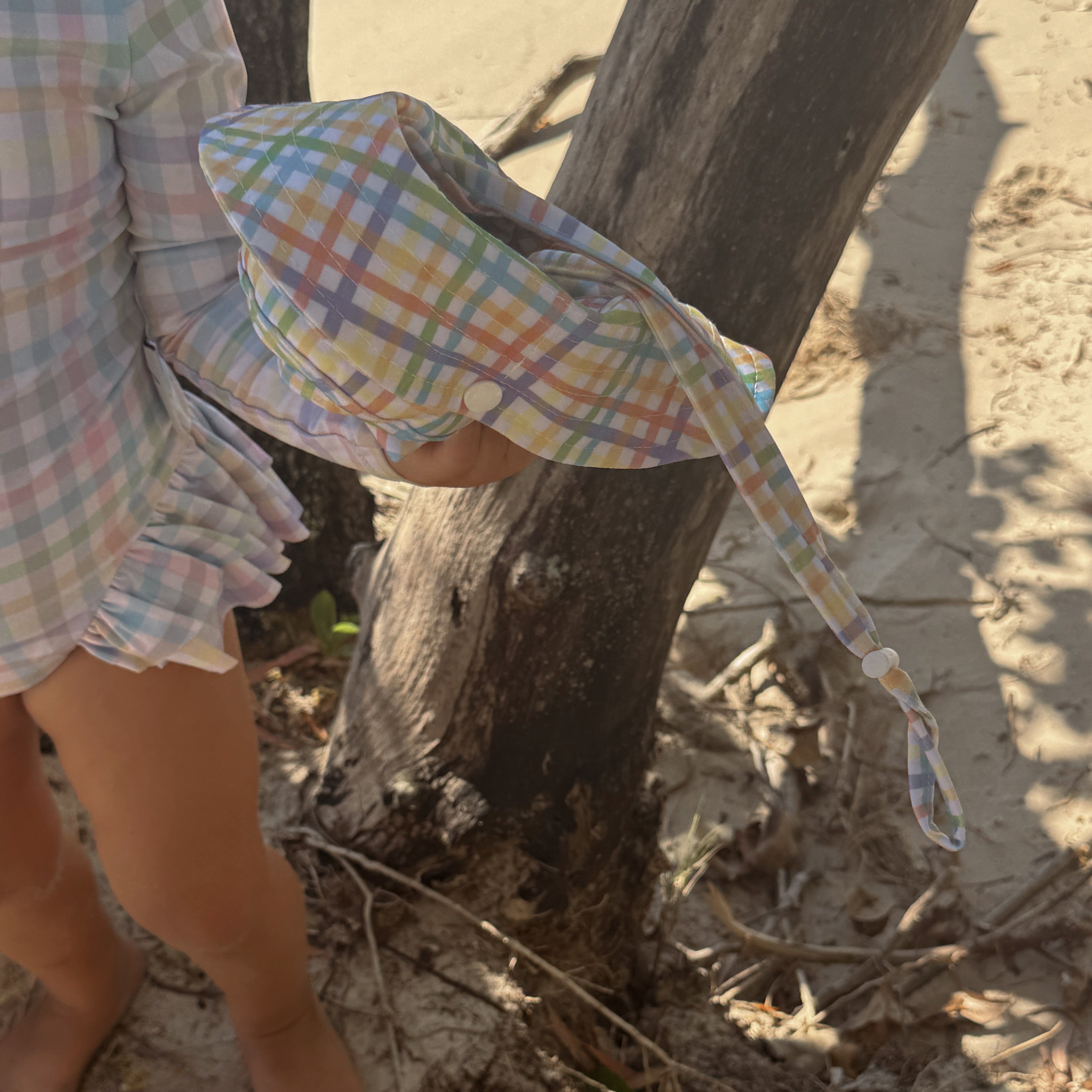 Child wearing a colorful checkered hat and dress standing on a sandy beach.