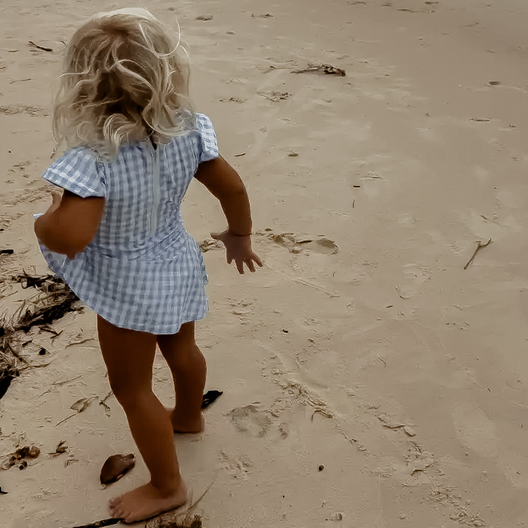 Child in a blue checkered swimsuit dress walking on a sandy beach.
