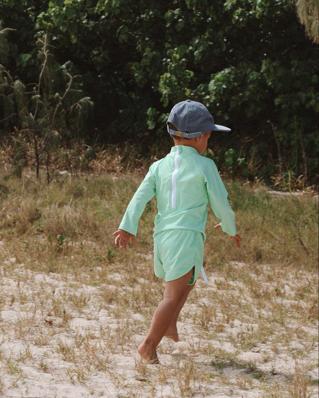 Boy in a light green swim shirt and cap walking on a sandy path with greenery in the background