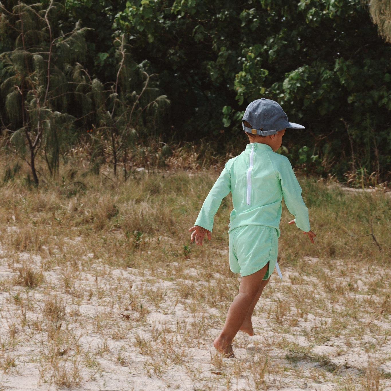 Boy in a light green swim shirt and cap walking on a sandy path with greenery in the background