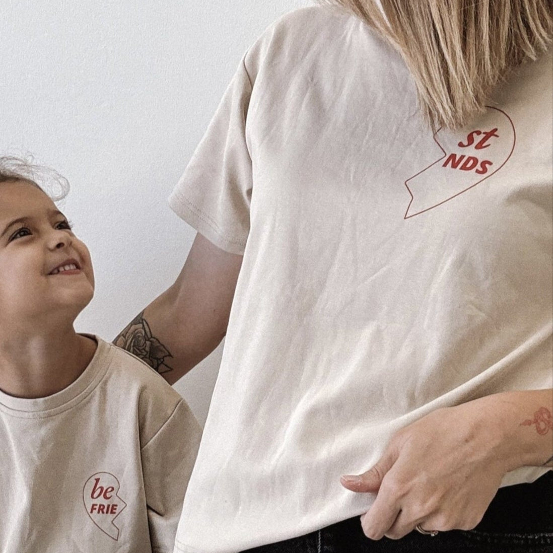 Woman and child wearing matching beige t-shirts with a logo, sitting on a wooden stool.