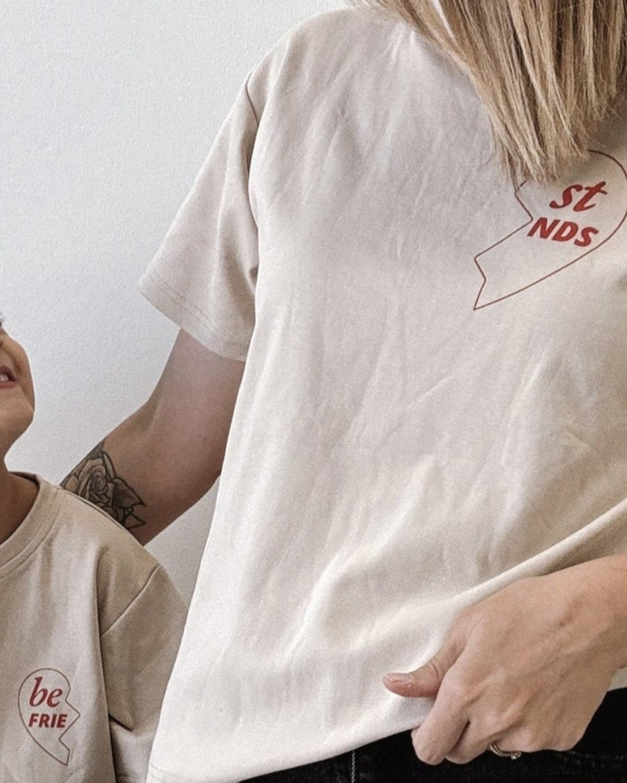 Woman and child wearing matching beige t-shirts with a logo, sitting on a wooden stool.