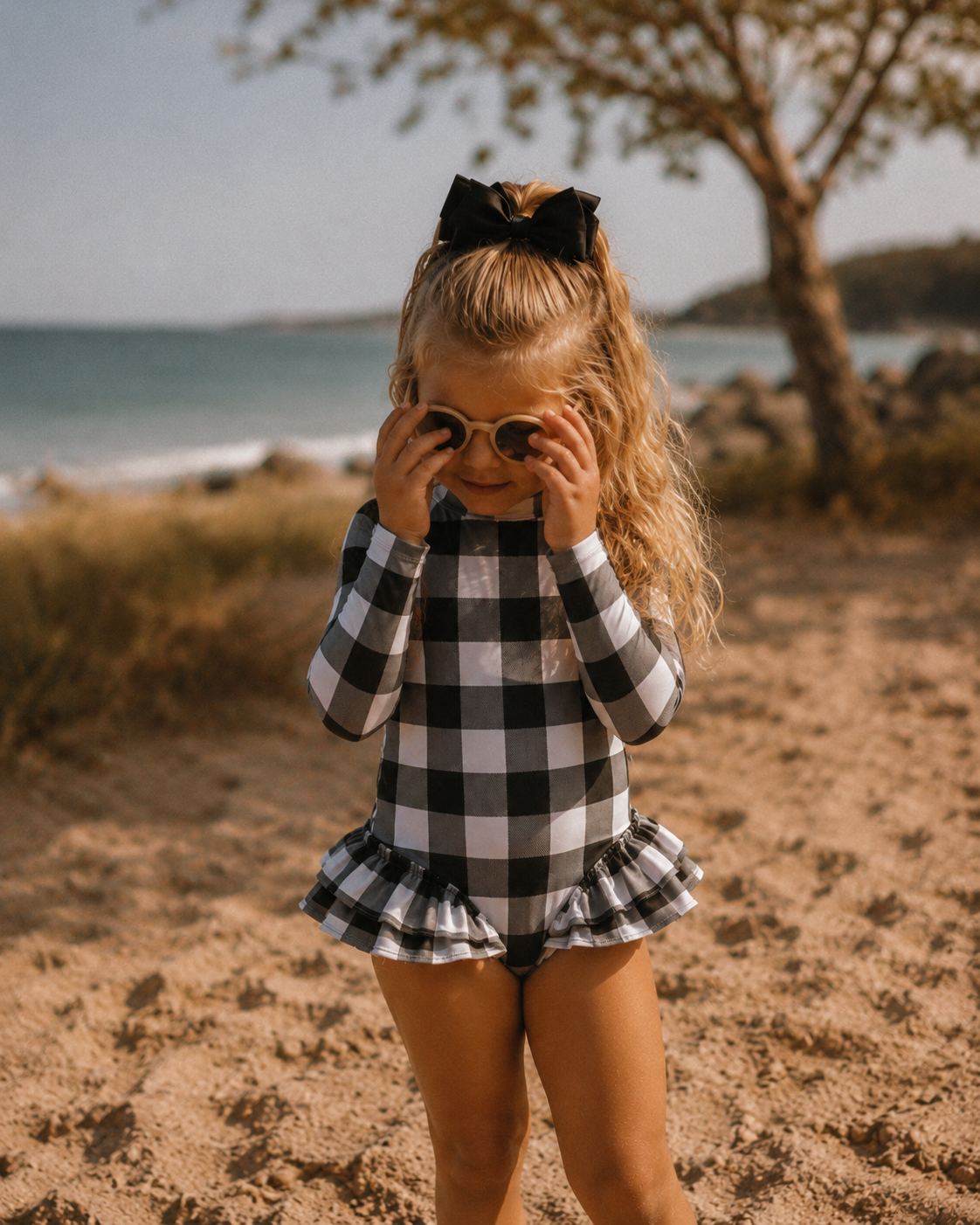 A young girl with a black bow in her hair stands on a sandy beach wearing a black and white buffalo plaid long-sleeve swimsuit with a ruffled hem. She is holding her sunglasses up to her eyes.
