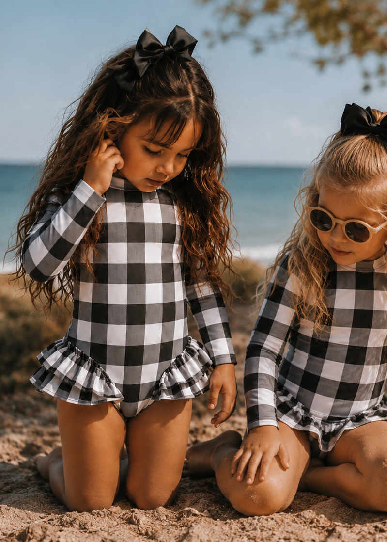 Two young girls kneeling on a sandy beach wearing matching black and white buffalo plaid long-sleeve swim dresses. One girl has dark hair with a black bow and the other has blonde hair with sunglasses and a black bow.