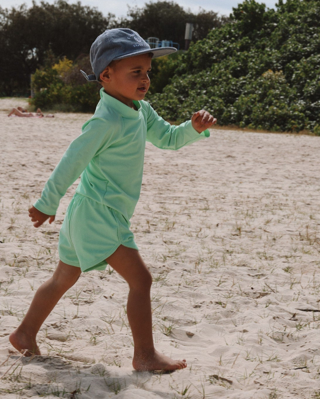 Child in a light green outfit and cap walking on sandy ground with greenery in the background