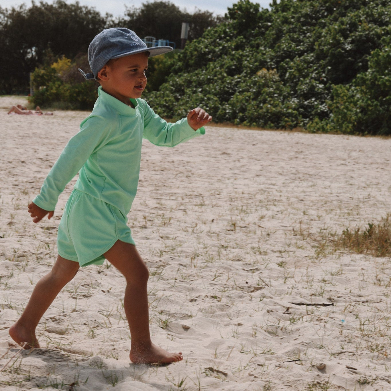 Child in a light green outfit and cap walking on sandy ground with greenery in the background