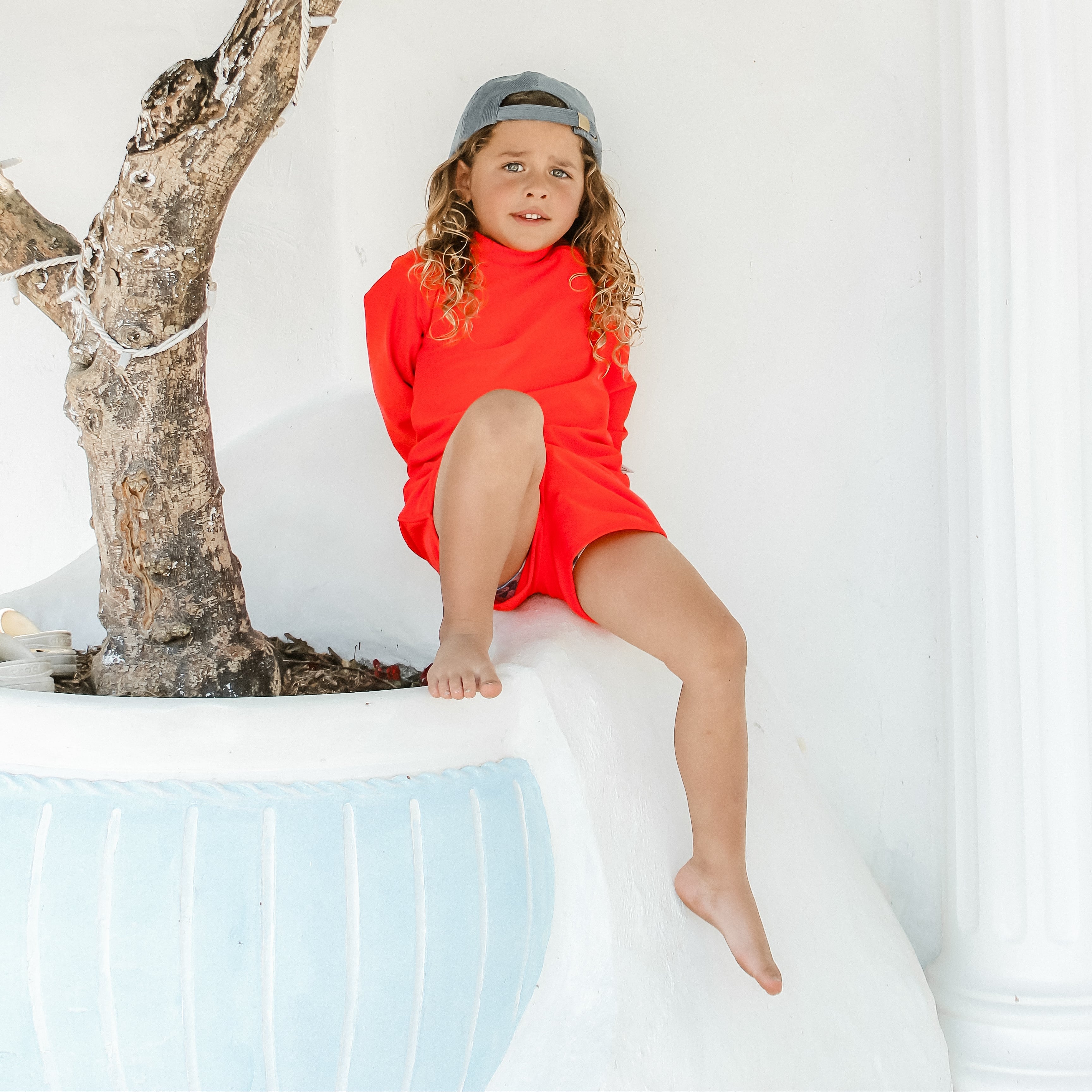 Child in a bright hi vis  red rash suit swim set sitting on a white ledge with a tree and white wall in the background