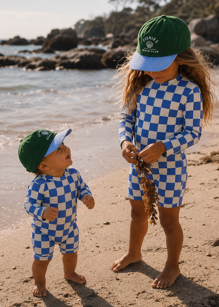 Two young children standing on a sandy beach wearing matching blue and white checkered long-sleeve swimsuits and green caps with light blue brims, with the ocean and rocks in the background.