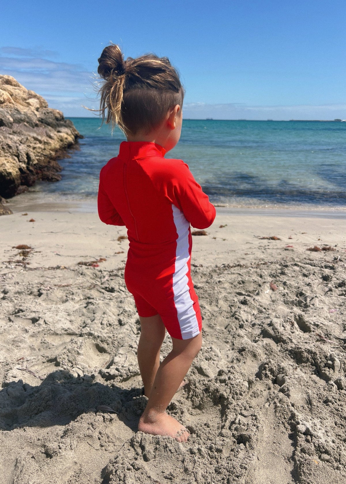 A child wearing a red and white long-sleeve swim rash guard and matching swim shorts stands on a sandy beach facing the ocean.