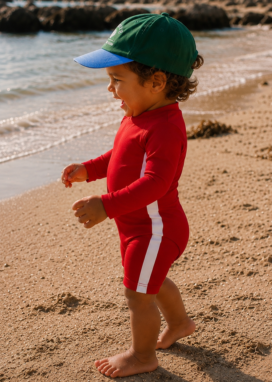 A toddler walking barefoot on a sandy beach wearing a red long-sleeve one-piece swimsuit with a white side stripe, a green and blue baseball cap, and a wetsuit.