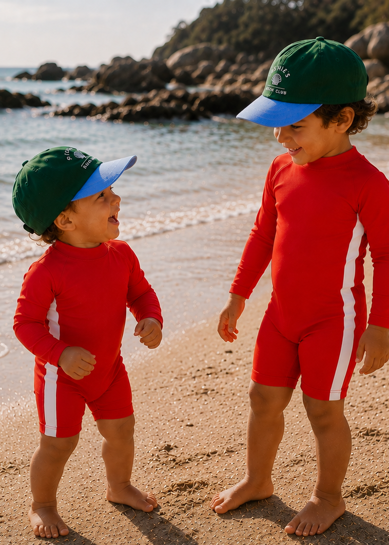 Two young children standing on a sandy beach wearing matching red long-sleeve swim suits with white side panels and green swim caps with blue visors, with the ocean and rocks in the background.