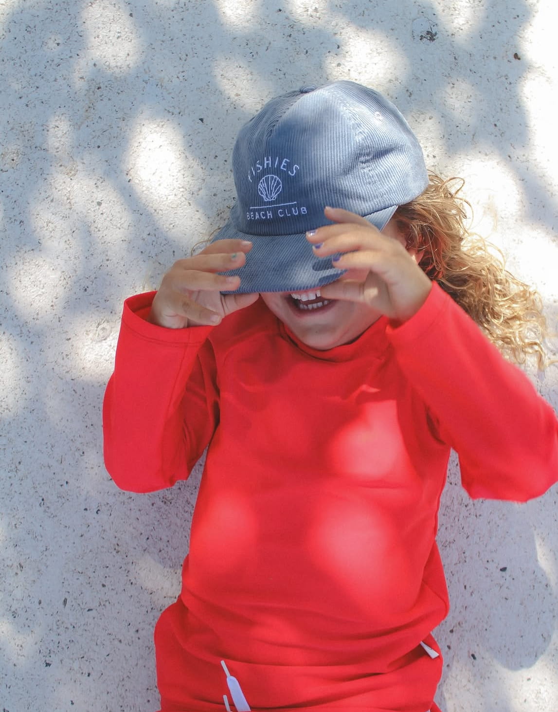 Child wearing a blue cap and red shirt, standing against a textured wall.