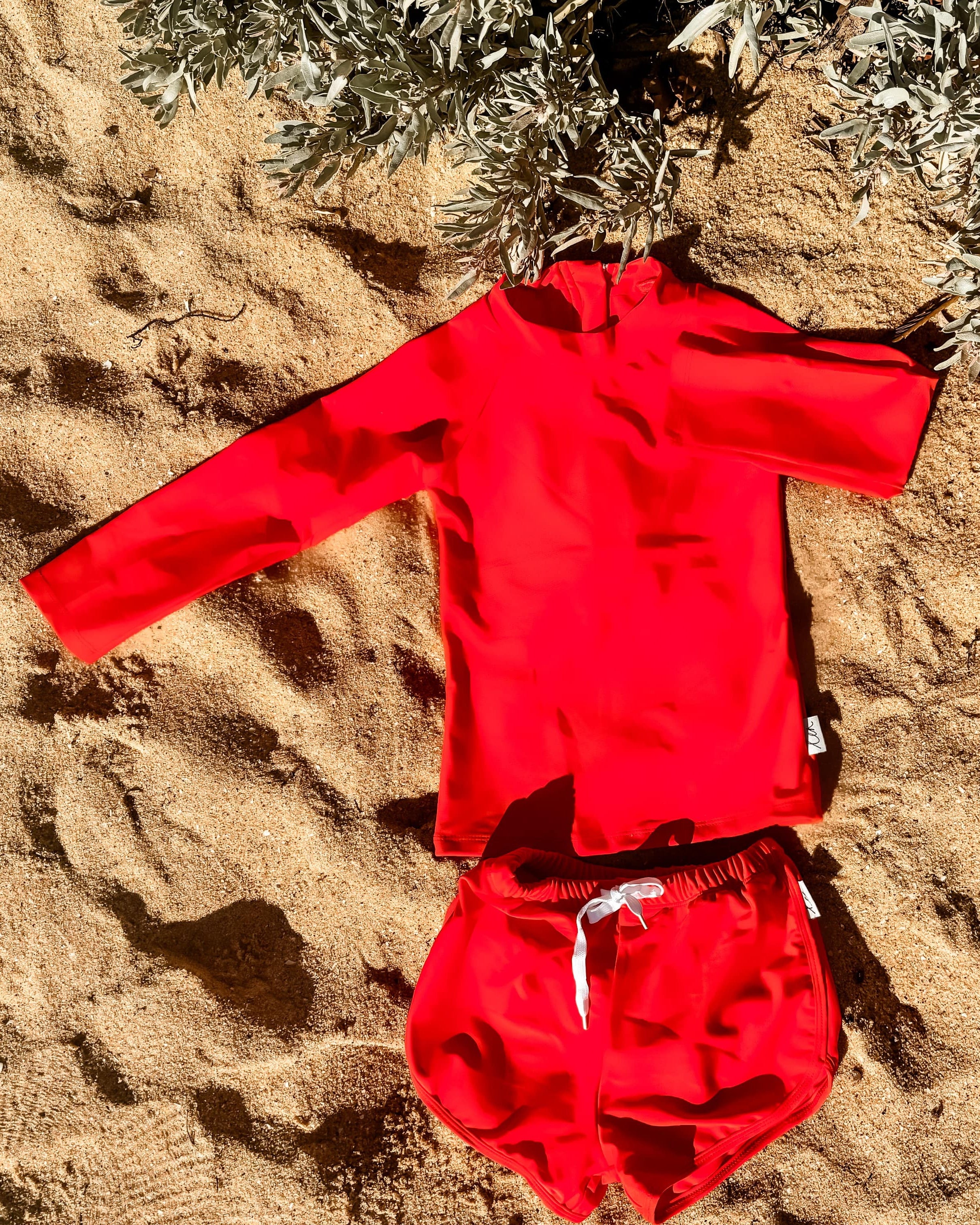 Red children's swimsuit set on a sandy surface with plants in the background
