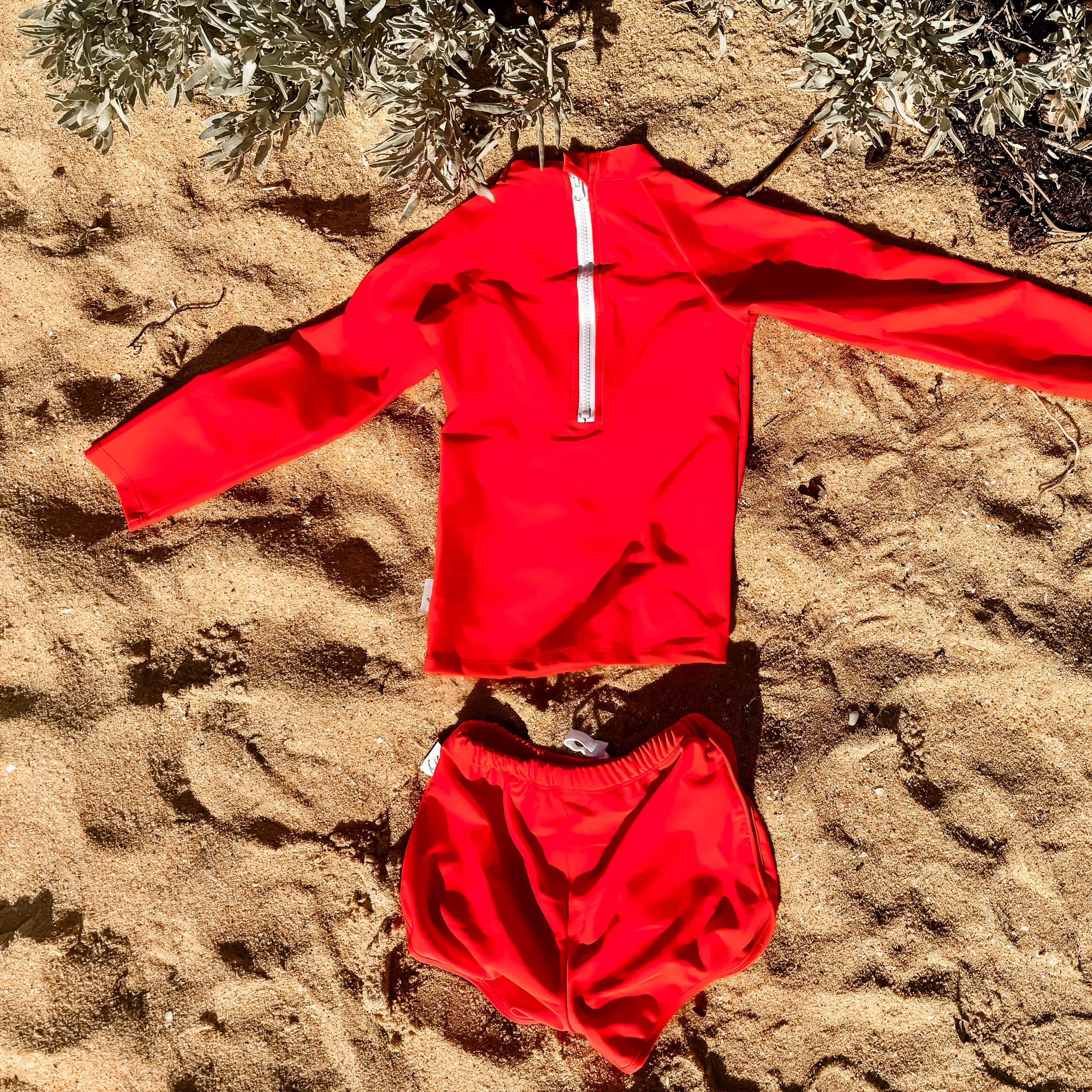 bright swimsuit for boys in a Red outfit laid out on sandy ground with plants in the background