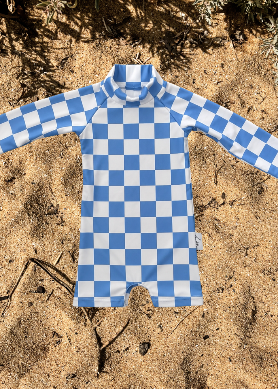 A long-sleeve baby romper with a blue and white checkerboard pattern, laid flat on sandy ground with some dry grass in the background.