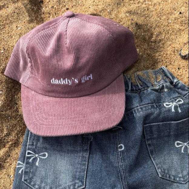 Pink cap with 'daddy's girl' text on sand next to blue jeans with bow details

