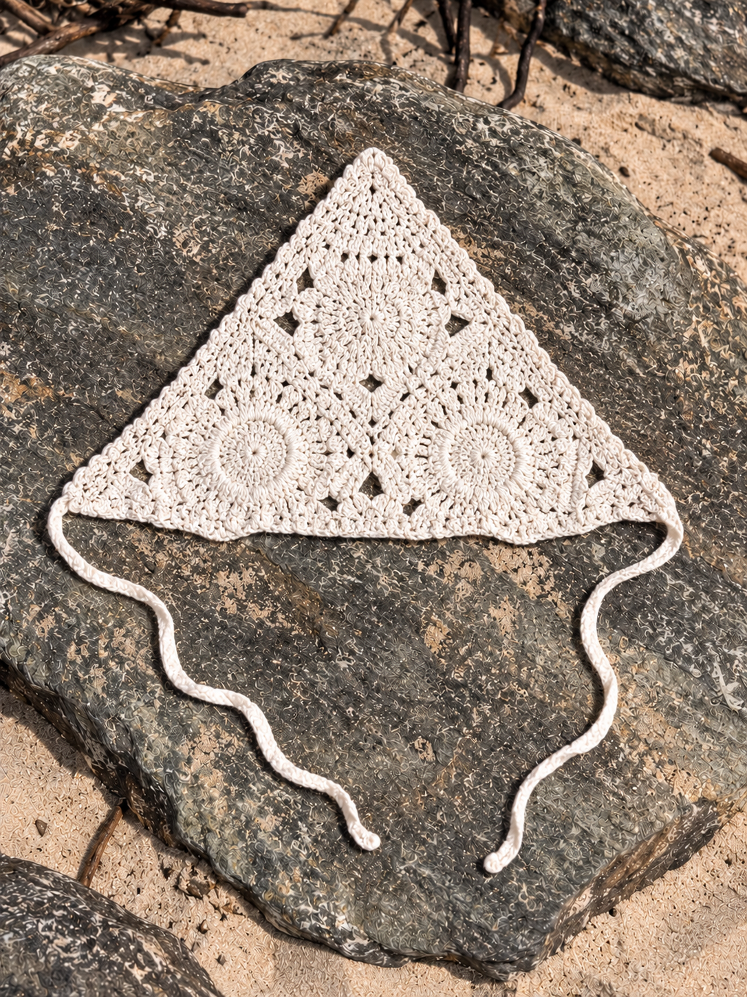 A handmade triangular crochet hat in off-white, laid flat on a large dark rock outdoors with sand visible around it.