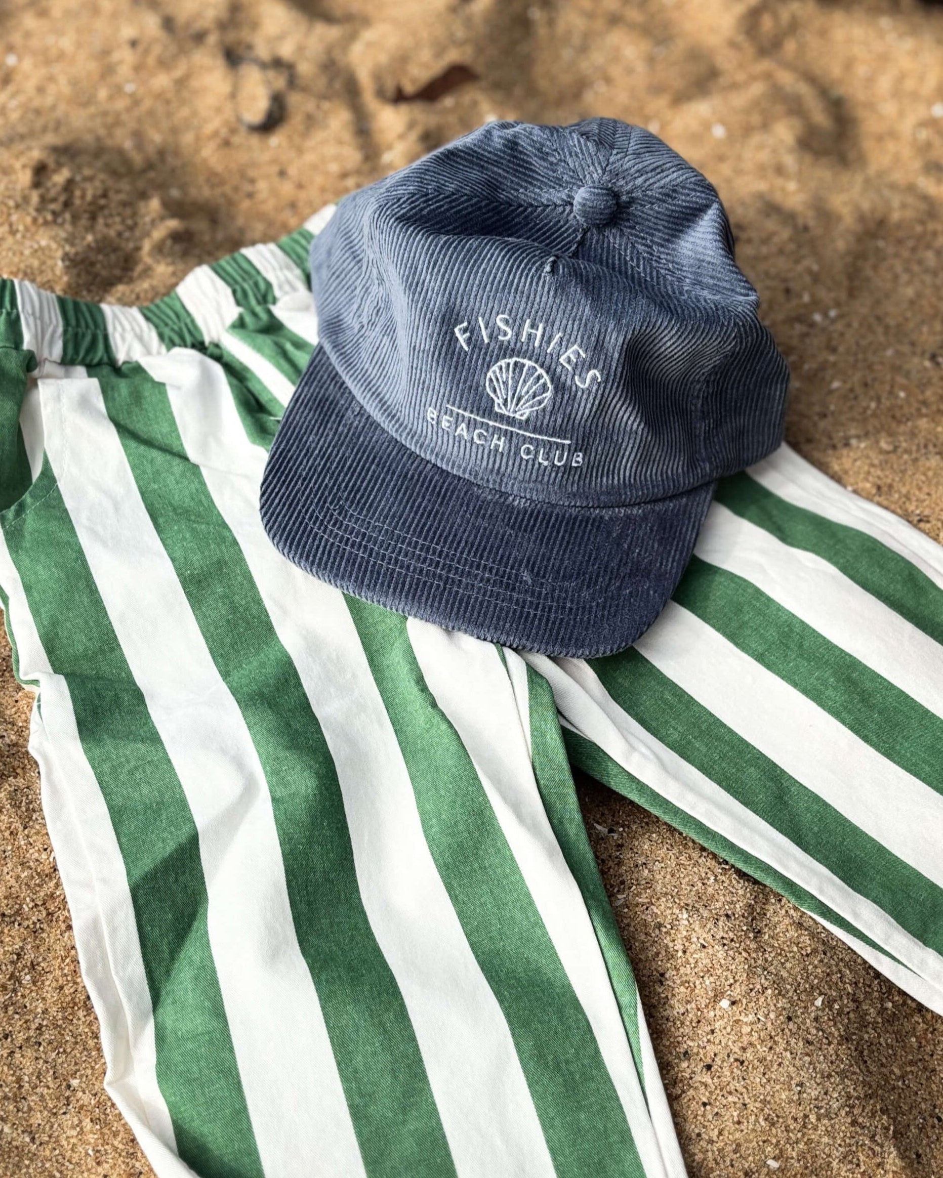 Green and white striped toddler boys summer pants with a blue cap on sandy ground
