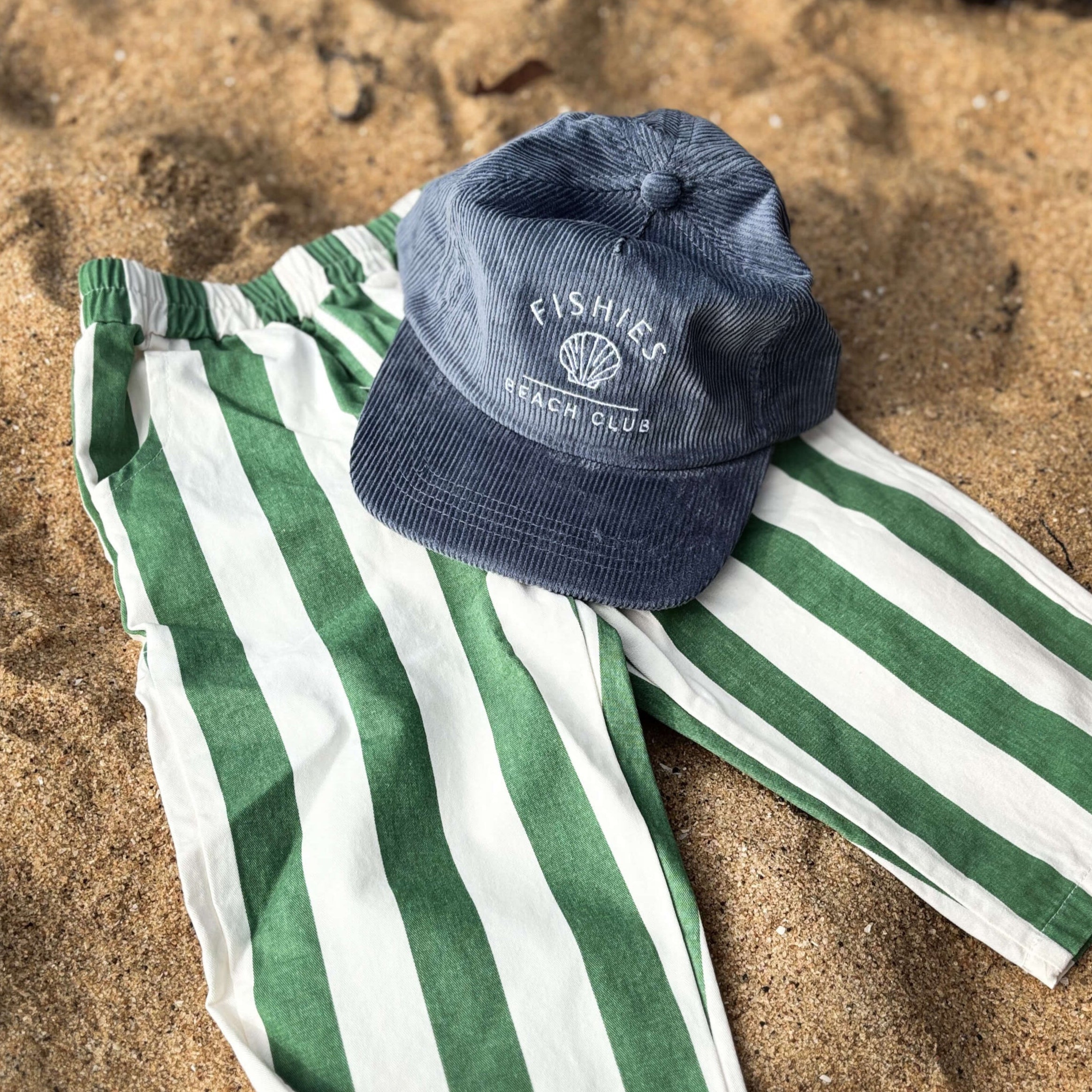 Green and white striped toddler boys summer pants with a blue cap on sandy ground
