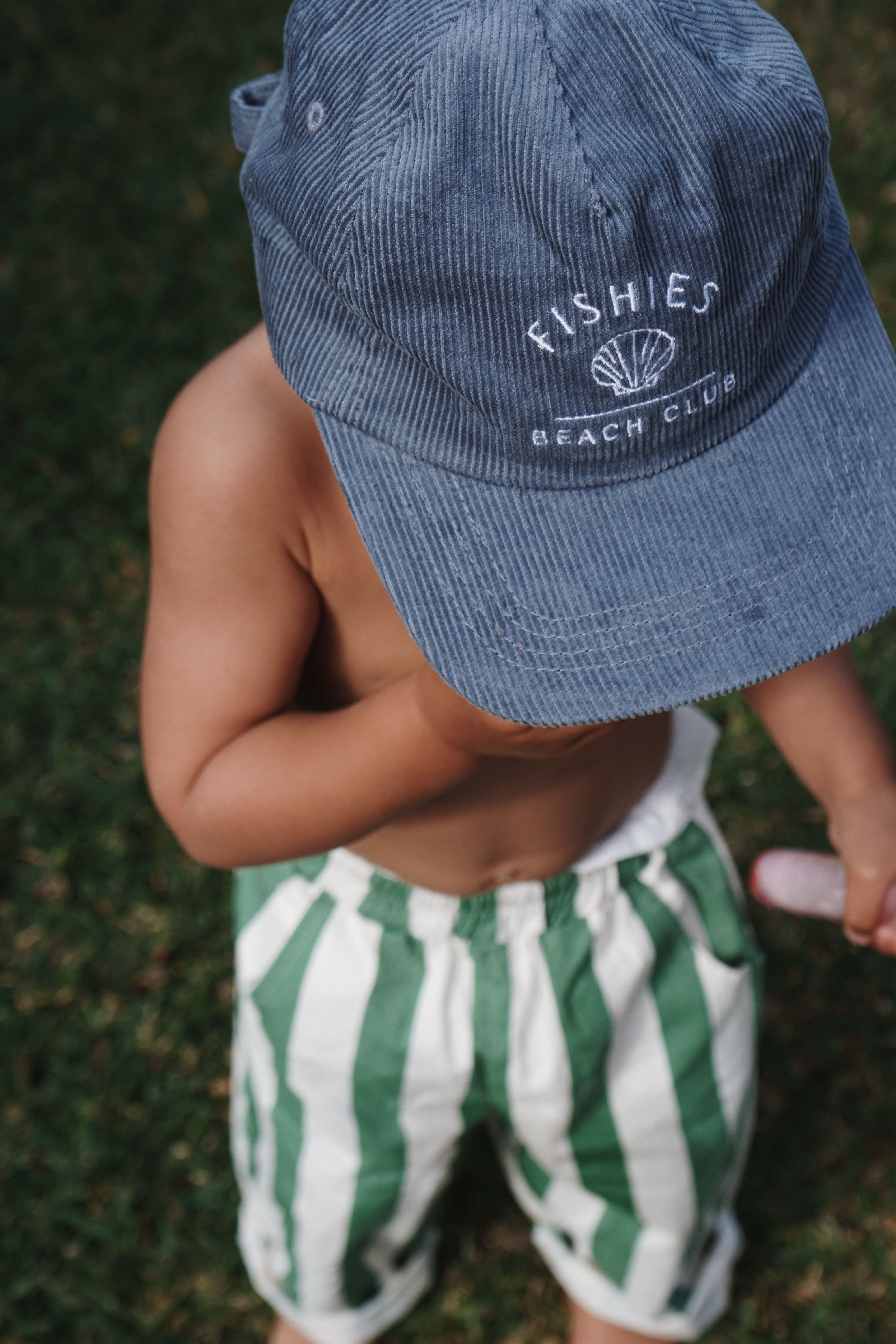 Child wearing a blue cap with 'Fishies Beach Club' logo and green and white striped shorts on grass.