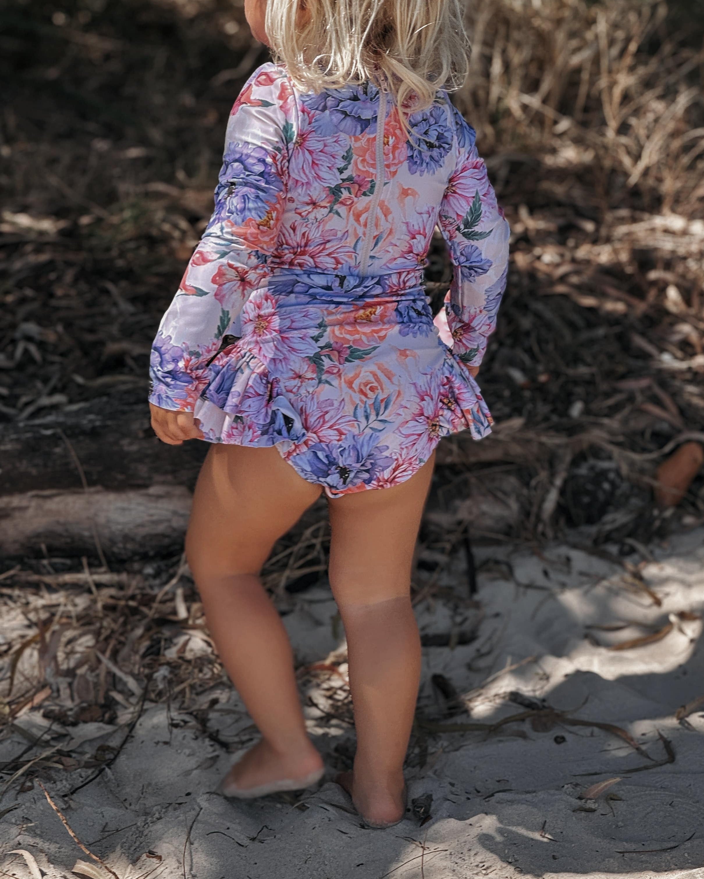 Child in a floral dress standing on a sandy beach with dry grass and twigs wearing a long sleeve upf50 sun safe swimsuit with no toxins and nappy change snapsaround.
