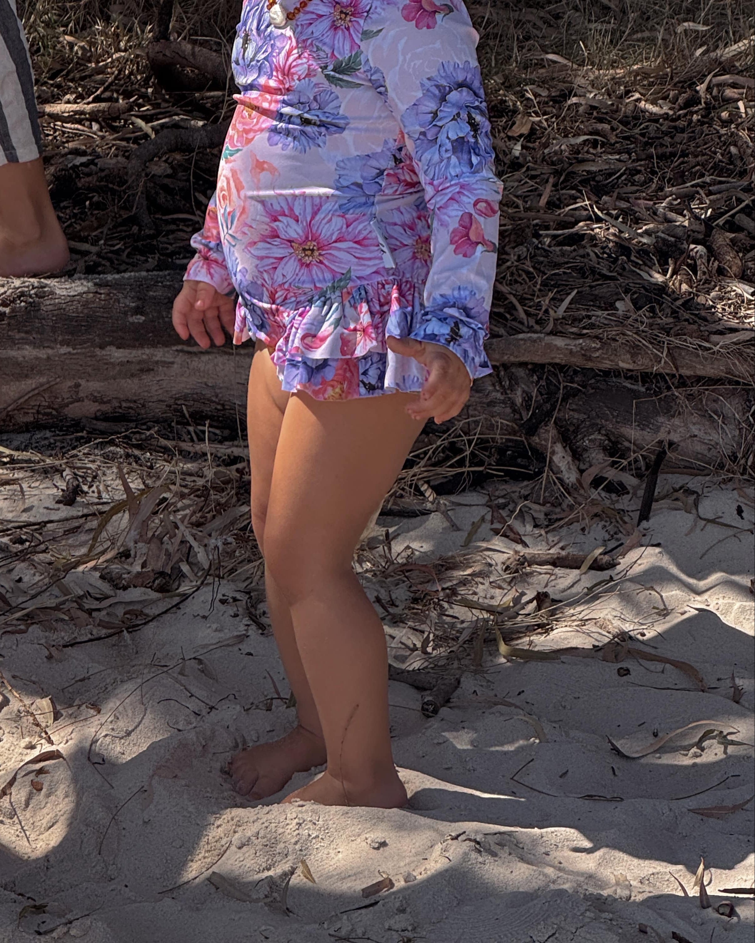 baby girl wearing a colorful floral \wimsuits\s standing on sandy ground with another person partially visible.