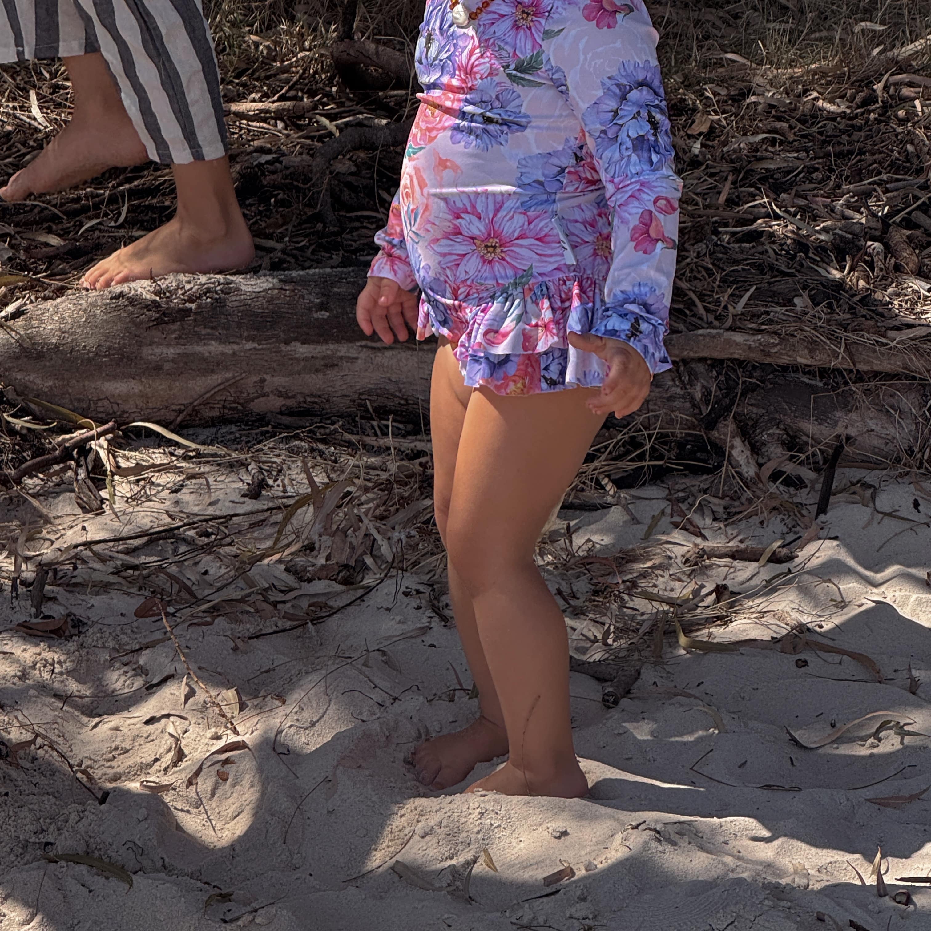 baby girl wearing a colorful floral \wimsuits\s standing on sandy ground with another person partially visible.