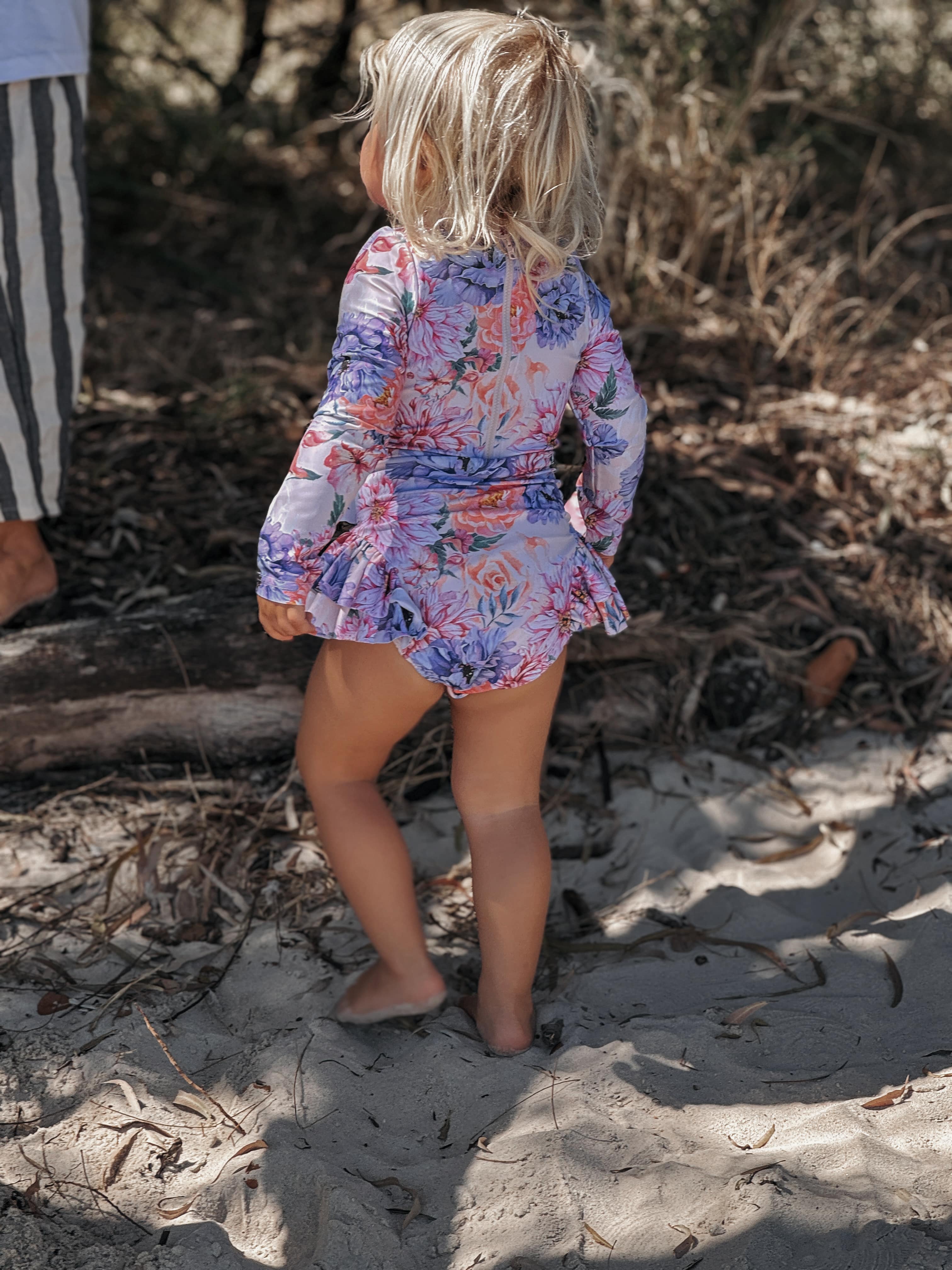 Child in a floral dress standing on a sandy beach with dry grass and twigs around.