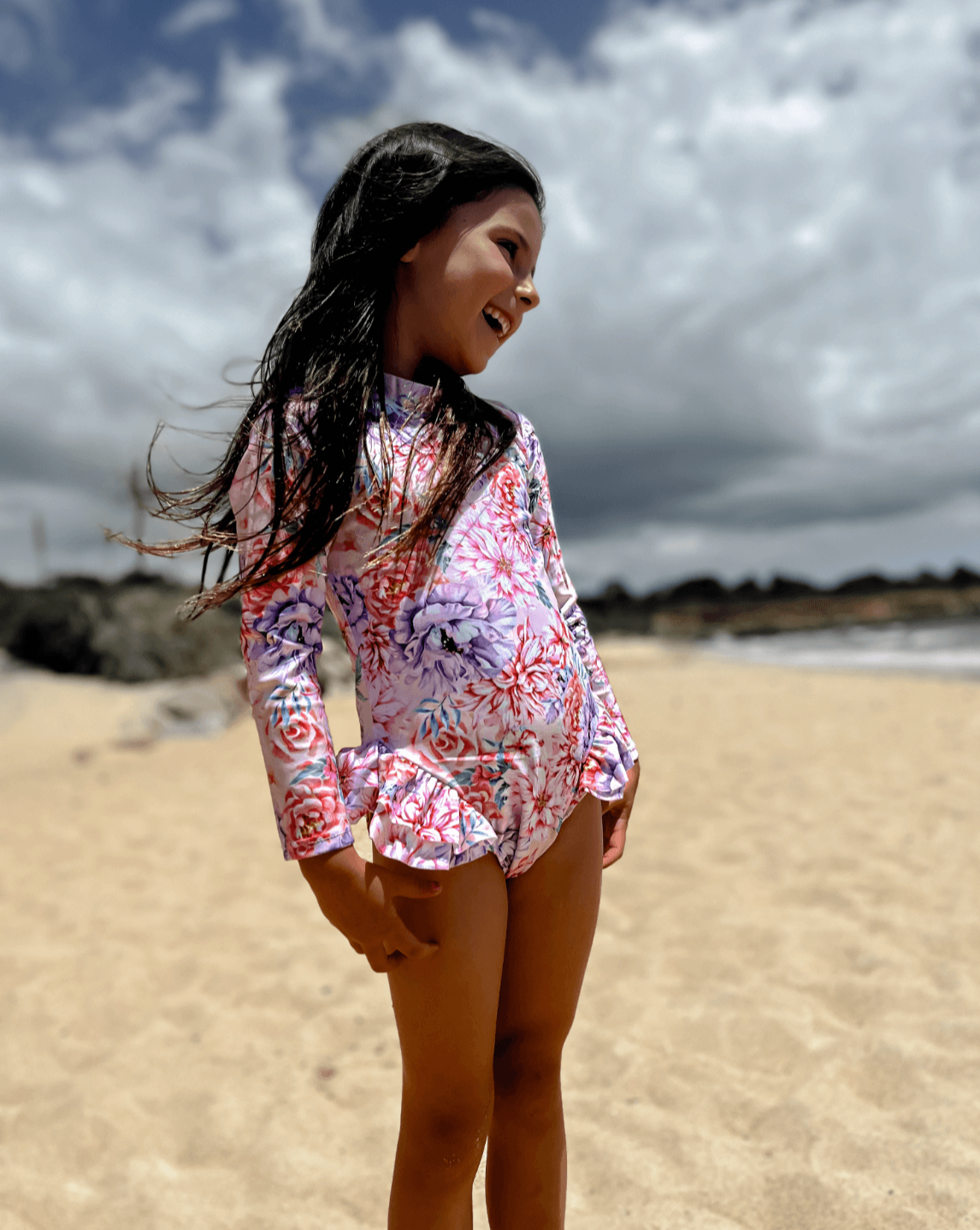 Girl at beach wearing a premium long sleeve swimsuit that has nappy/change snaps in a pink and purple hydrangea and peony floral print.