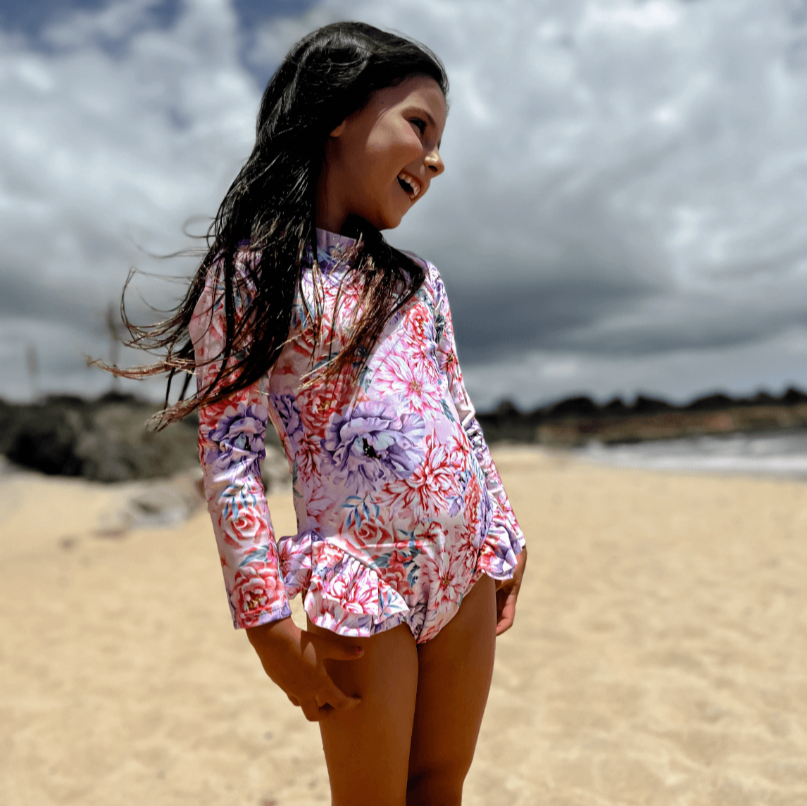 Girl at beach wearing a premium long sleeve swimsuit that has nappy/change snaps in a pink and purple hydrangea and peony floral print.