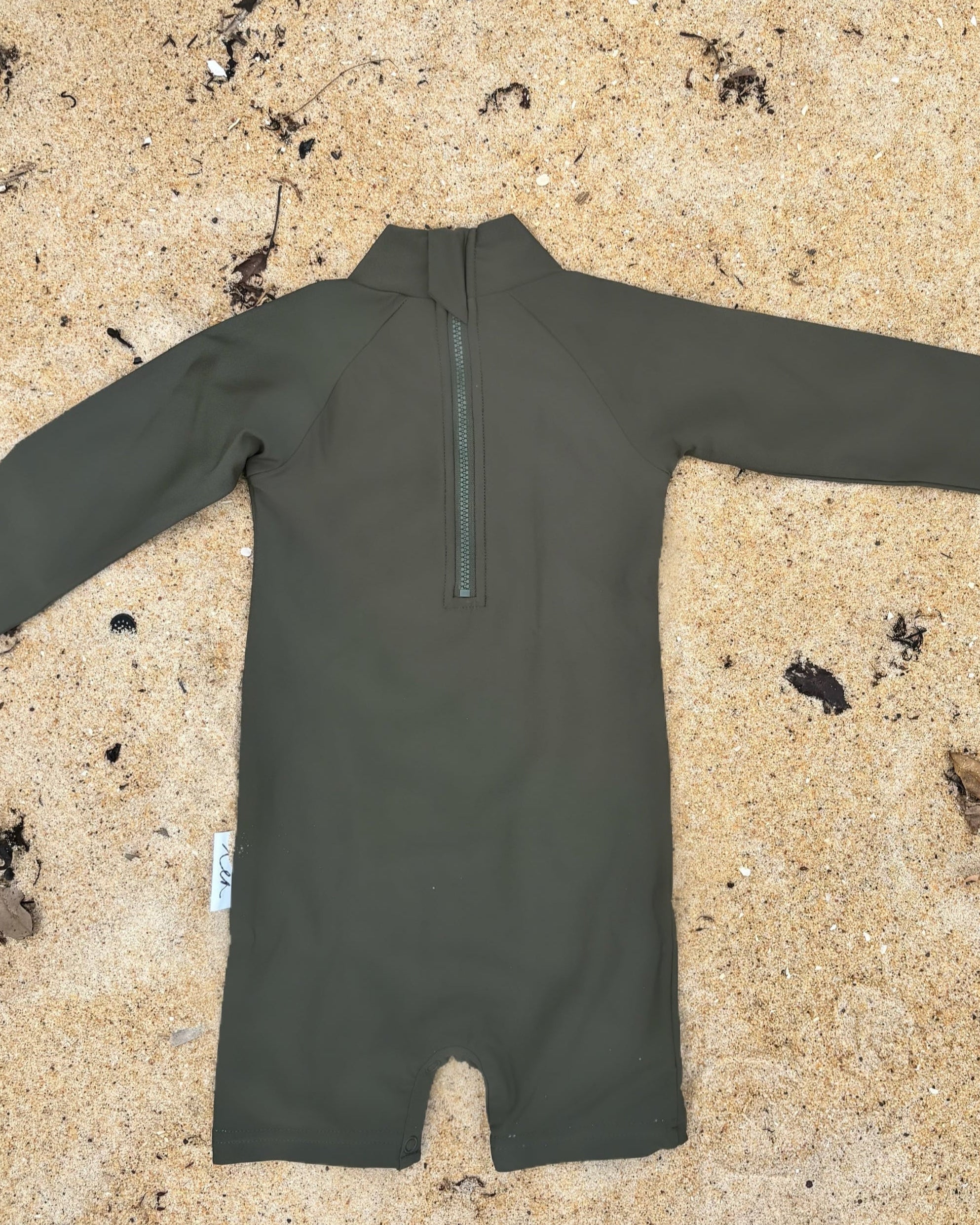 Dark Green infantswimsuit on a sandy surface with plants in the background