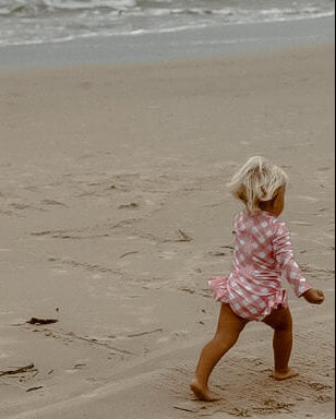 Child in a pink checkered outfit running on a sandy beach with waves in the background