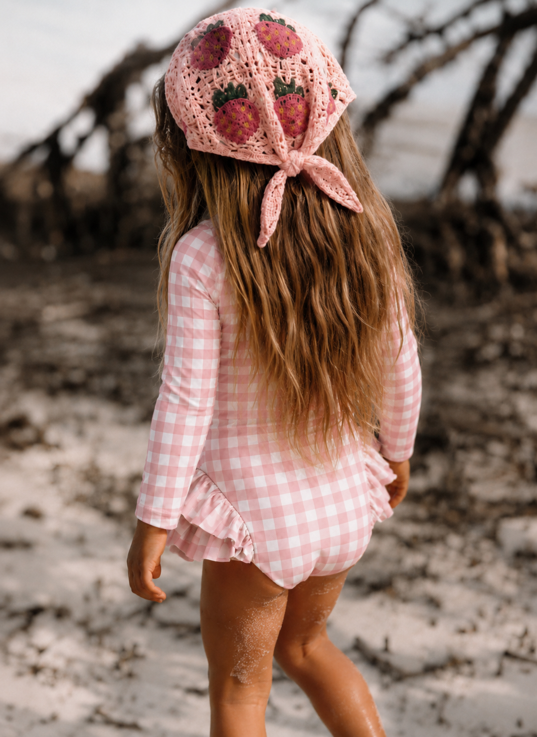 A young girl seen from behind on a beach, wearing a pink and white gingham long-sleeve romper and a pink crochet sun hat with a strawberry pattern.