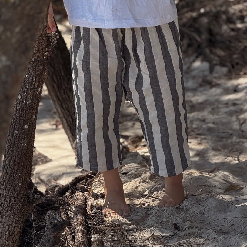 Boys beachwear light weigh pant wearing striped pants standing on a sandy ground with dry vegetation.